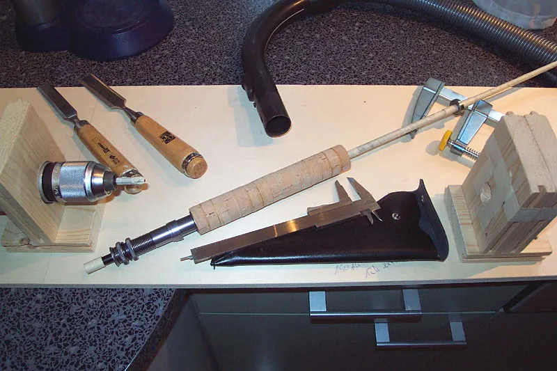 Preparations done for leveling and sanding - The blank is covered with masking tape and a bearing is placed on a masking tape bushing to avoid any harm to the blank. At the rear end of the blank I have placed a reinforcement wrap of masking tape. Tools from left to right: two sharp chisels, head of drilling machine with prepared chuck to fit the butt end of the blank. Caliper for measurings during the turning process and on the right the stand for the bearing and the necessary C-clamp behind it. In the background the end of the vacuum cleaner hose.